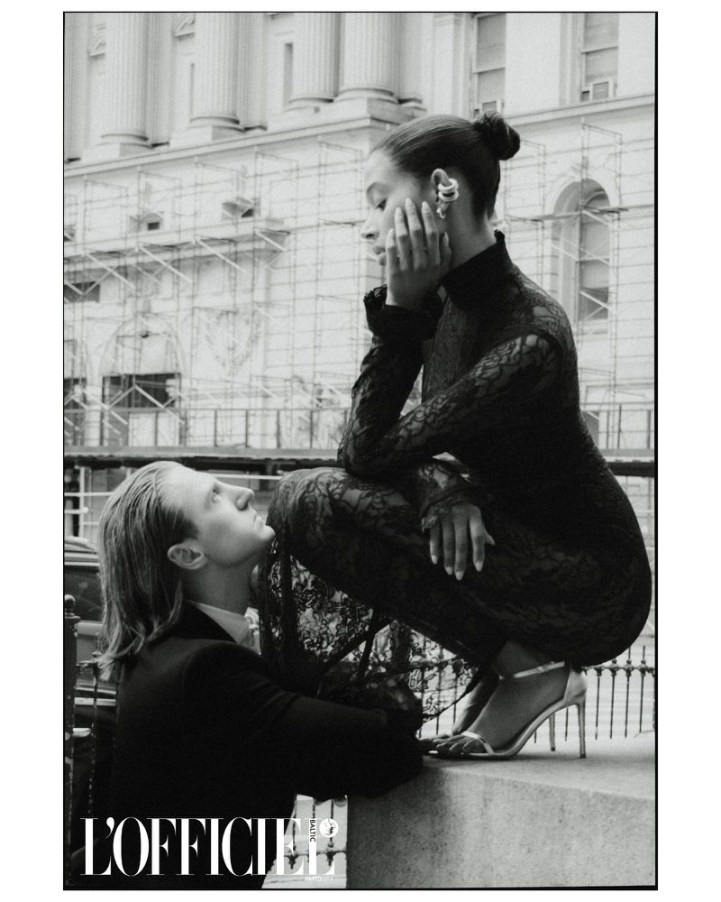 Black and white editorial fashion shot of a woman in a lace dress and silver heels posing with a man against a cityscape backdrop.