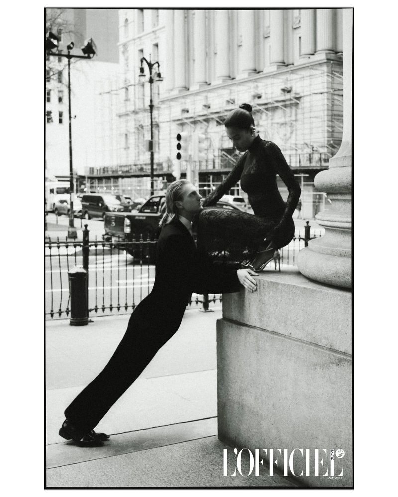 Black and white fashion shot of a man and woman posing against city architecture in structured clothing in an outdoor setting.