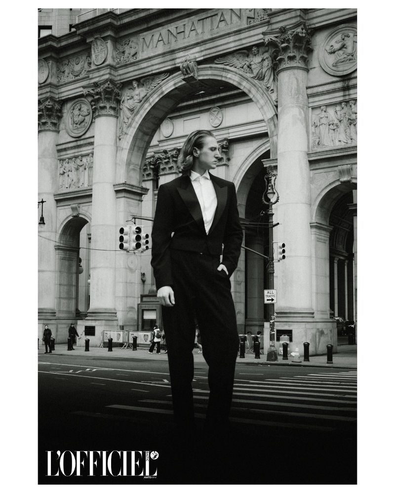 A long-haired man in a tuxedo poses on a city street in front of a grand archway with Manhattan inscribed on the architecture.