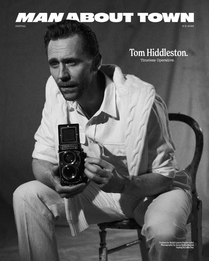 Actor Tom Hiddleston holding a vintage twin-lens reflex camera while seated on a chair in a monochrome portrait.