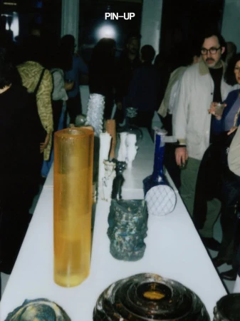 Candid event photograph showing a long white display table featuring assorted sculptural art objects, with attendees blurred in the background
