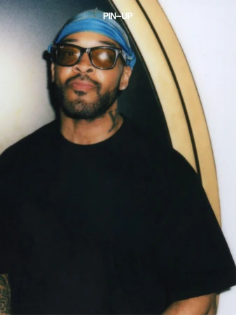 Black man wearing dark sunglasses and blue head covering with black t-shirt against dark background for PIN-UP Magazine