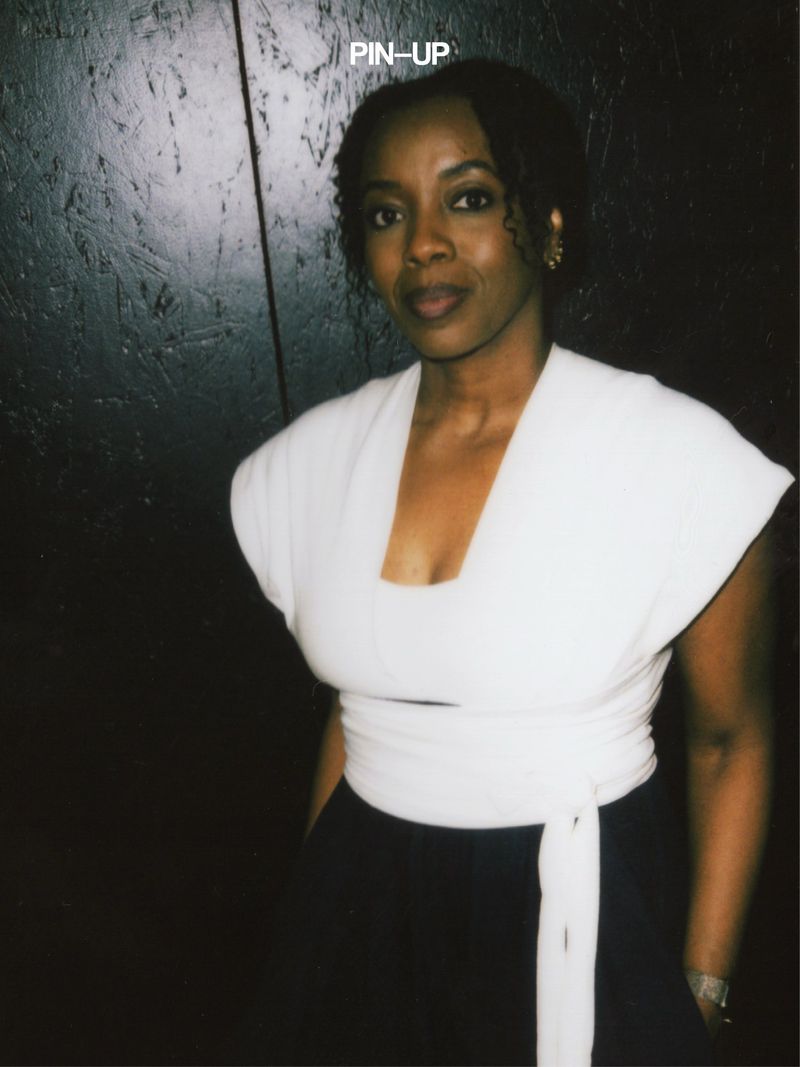 Portrait of a Black woman in a white wrap top against a dark paneled wall with PIN-UP text visible