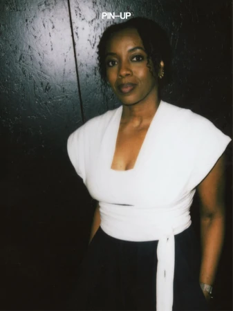 Portrait of a Black woman in a white wrap top against a dark paneled wall with PIN-UP text visible