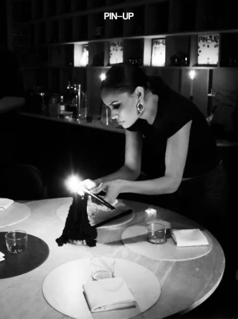Monochrome image of a woman leaning over a round dining table, lighting a candle in a dimly lit interior.