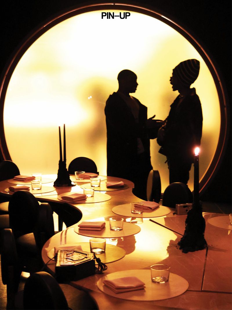Two silhouetted men talking in front of a large circular illuminated backdrop behind a set dining table.