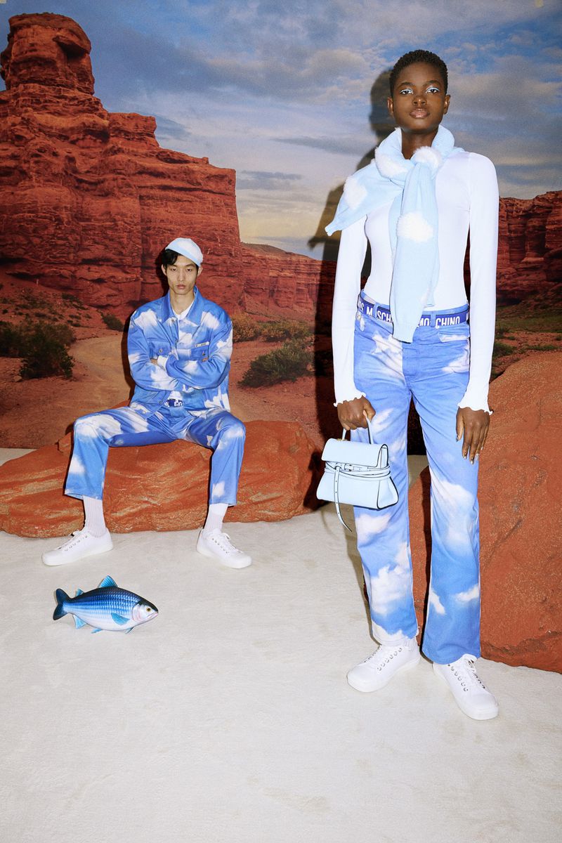Two fashion models wear coordinating sky-themed outfits against a desert backdrop in a studio shot featuring a toy fish and light blue bag.