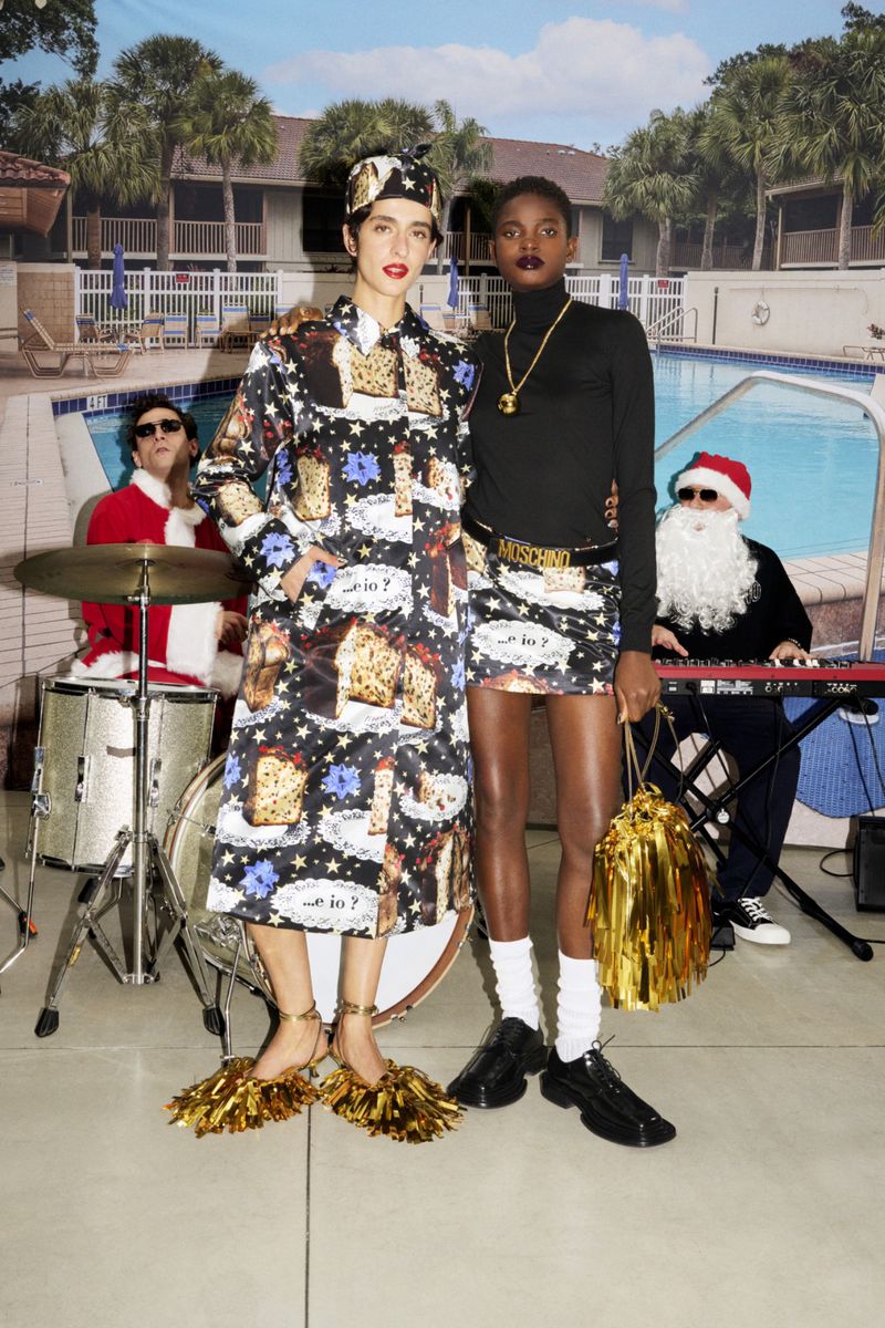 Two fashion models pose with a band in Santa Claus costumes by a pool, wearing cake-themed dresses with holiday and pop-art elements.
