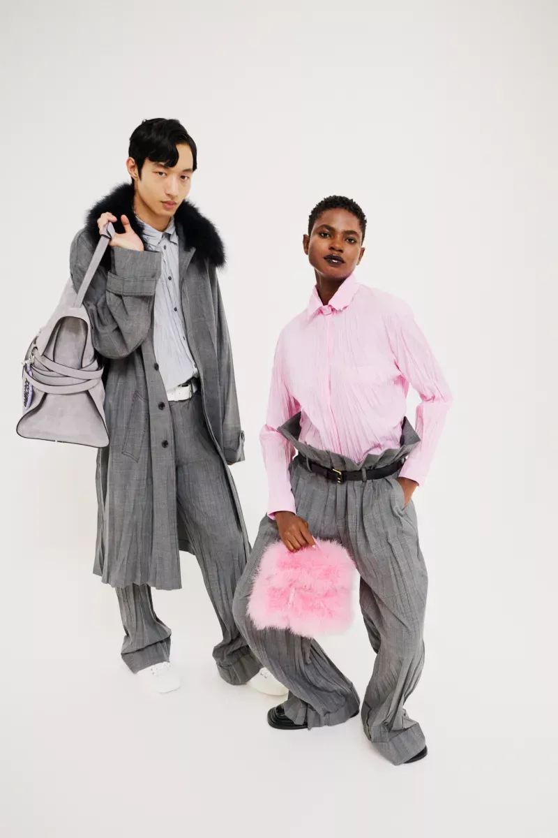 Two fashion models pose in a bright studio wearing grey pleated trousers and light-colored shirts, accessorized with bags.