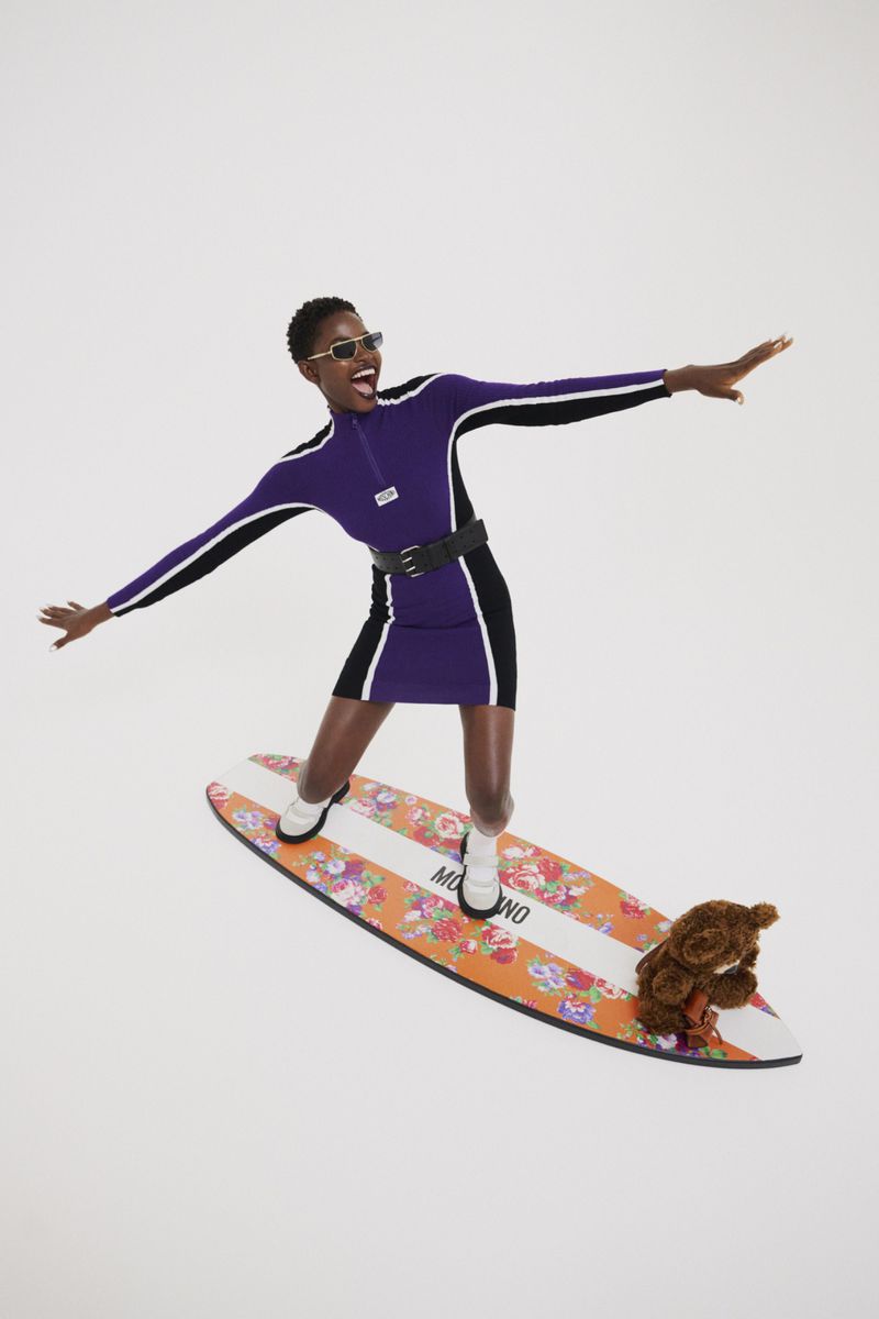 A black woman in a purple dress and sunglasses balances on a surfboard, smiling with arms outstretched, a teddy bear positioned near her.