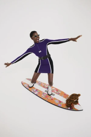A black woman in a purple dress and sunglasses balances on a surfboard, smiling with arms outstretched, a teddy bear positioned near her.