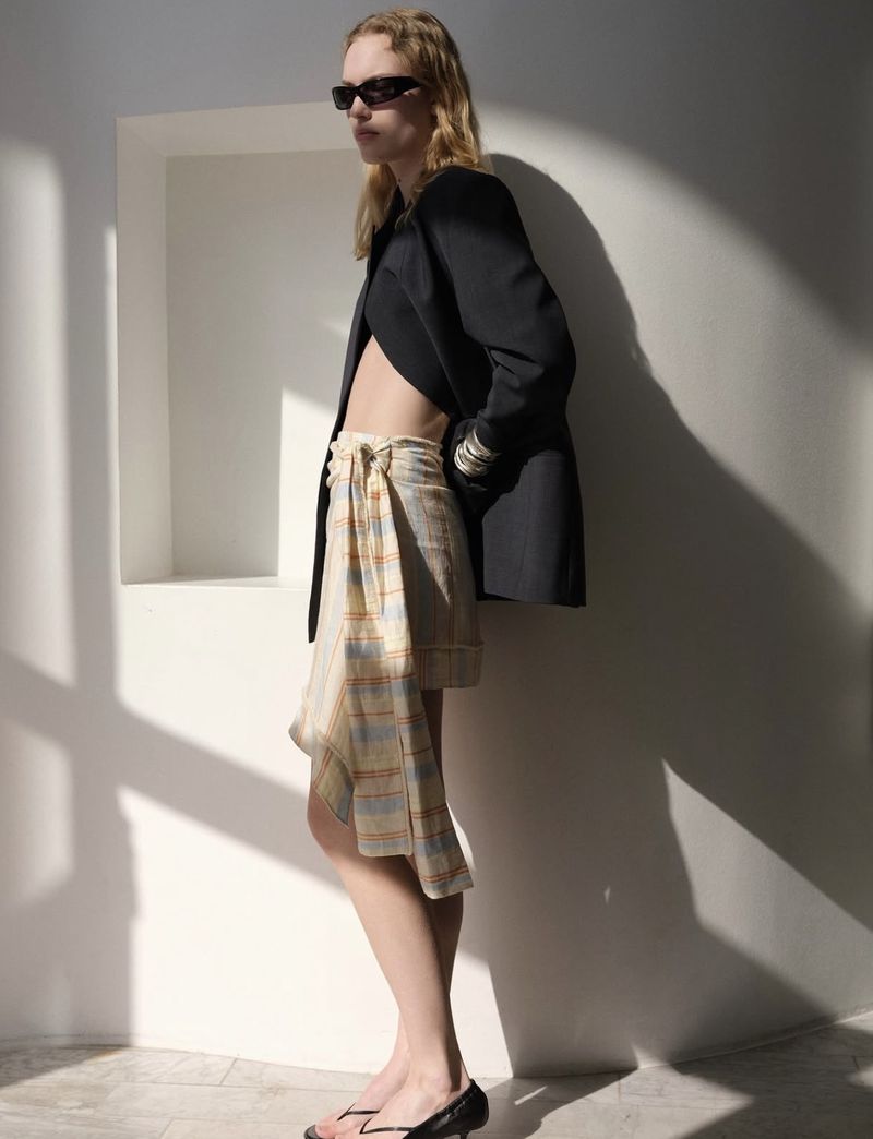 A fair-skinned woman poses in a black blazer and plaid skirt, wearing dark sunglasses against a white wall with soft window shadows.