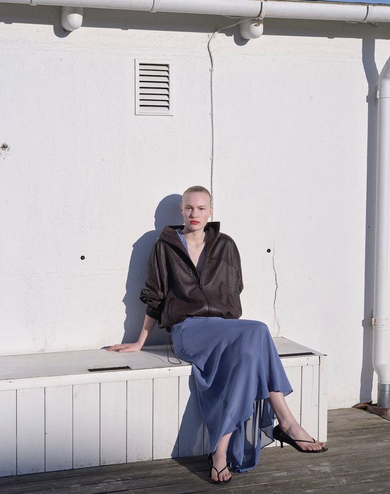 Model with short hair wearing a dark brown jacket and blue maxi skirt, seated against a white wall.