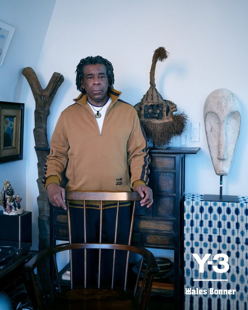 Portrait of adult male in casual fashion with short dark hair posing in room surrounded by tribal masks and objects with white wall.