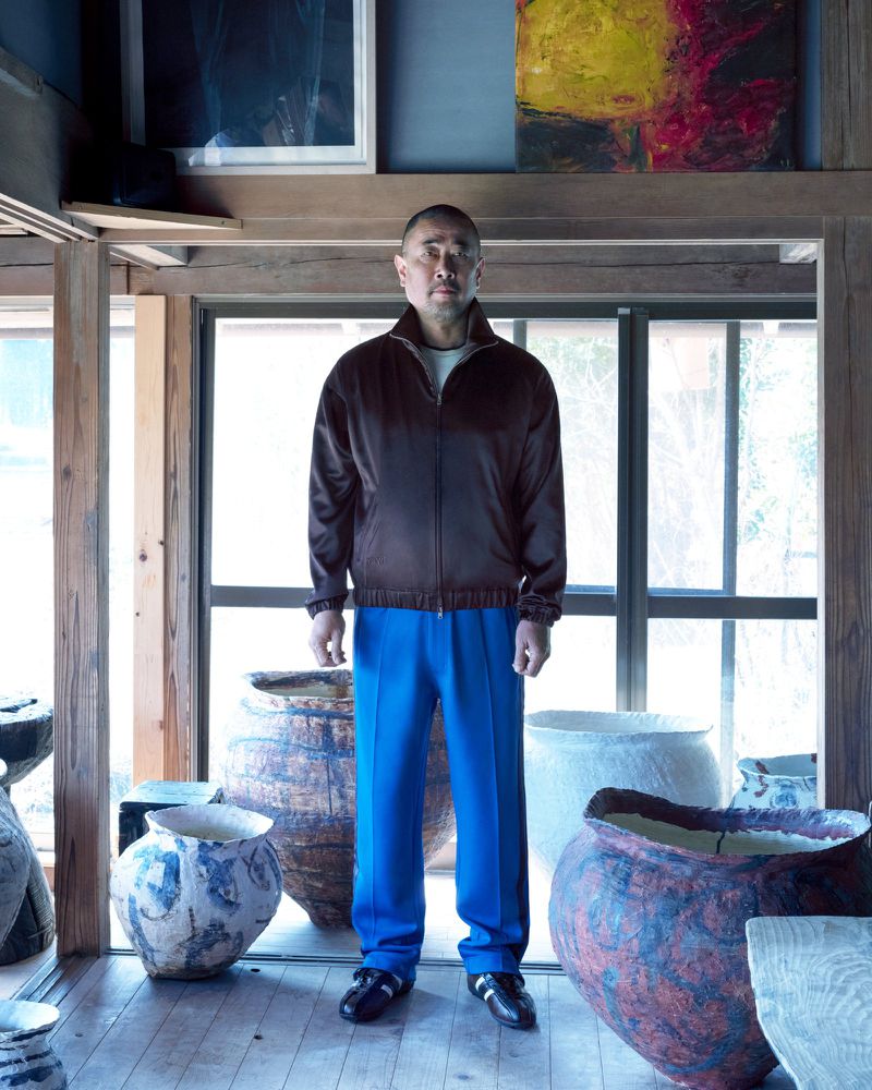 Man wearing a brown zip jacket and royal blue trousers standing indoors surrounded by ceramic vessels, photographed by Takashi Homma for Y-3 by Wales Bonner.