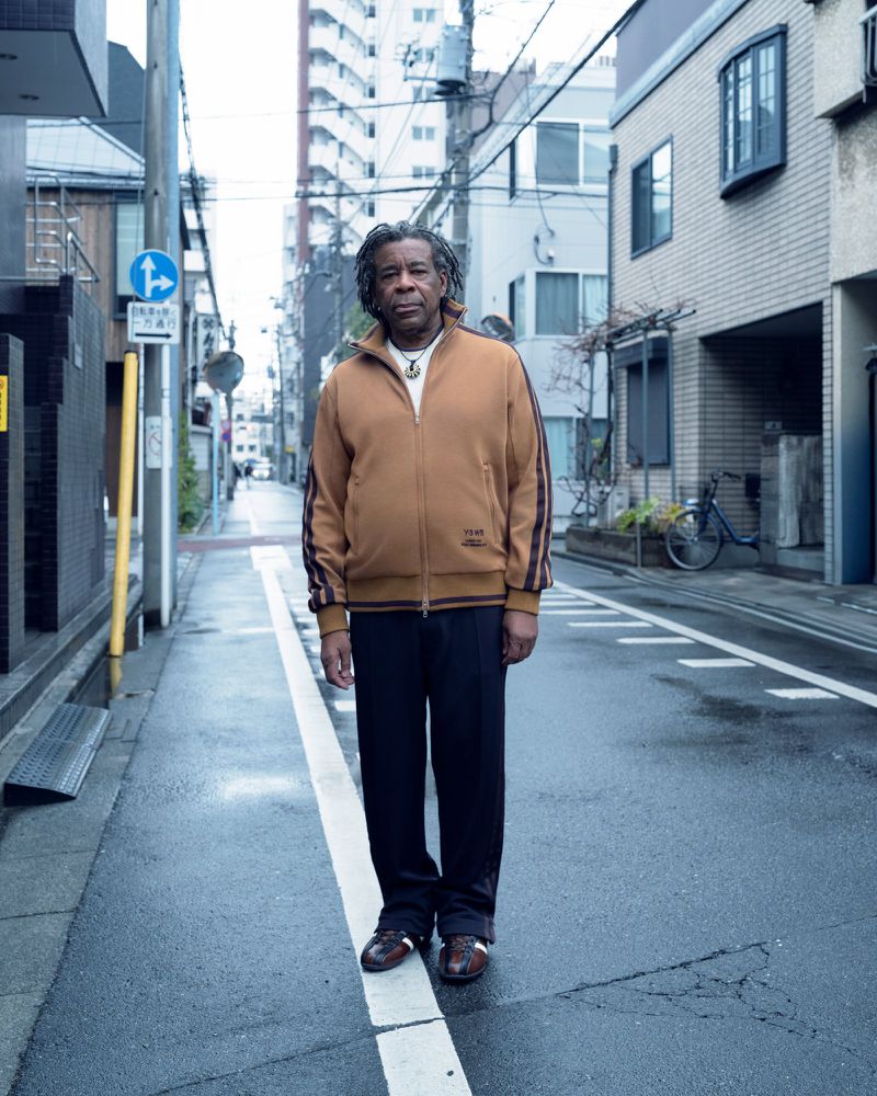 Man wearing a brown track jacket and dark trousers standing on a narrow urban street for Wales Bonner