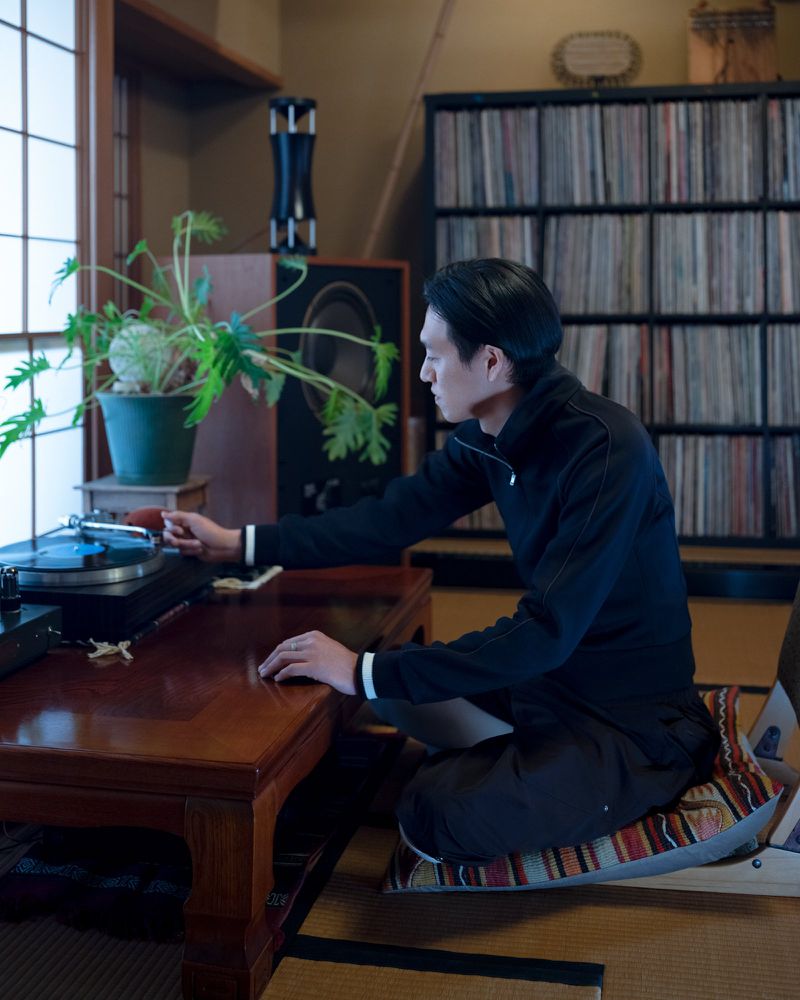 An Asian man seated on a tatami mat playing a record player inside a room with a record shelf and window light.
