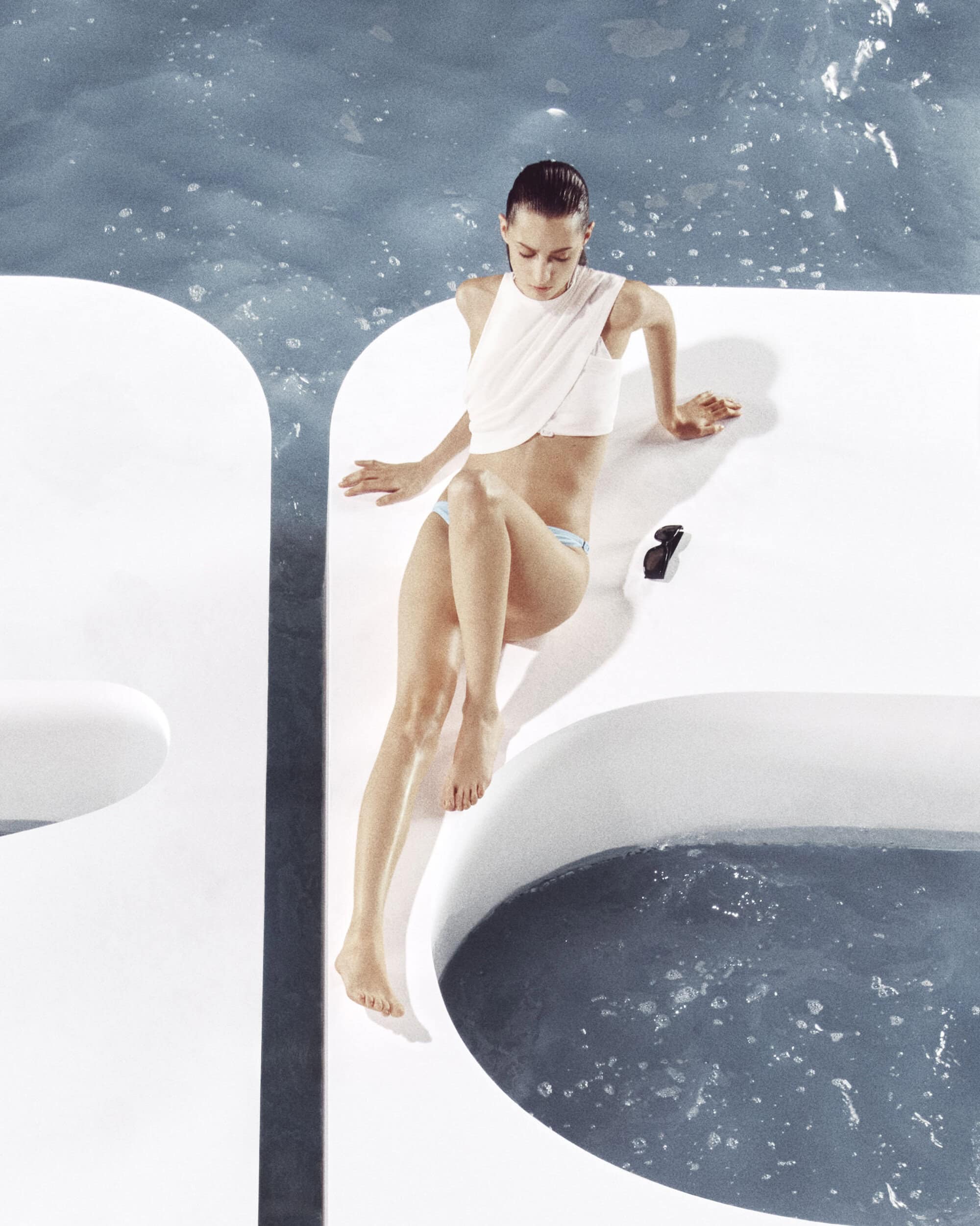 Model wearing a white top and blue bottoms seated on a white platform by water for Courreges