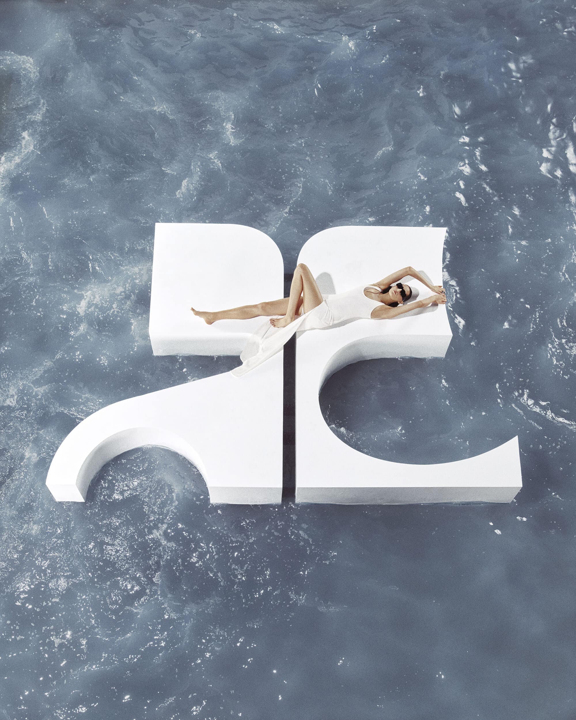 Model in white swimwear lounging on a large white Courreges logo platform floating on rippling blue water.