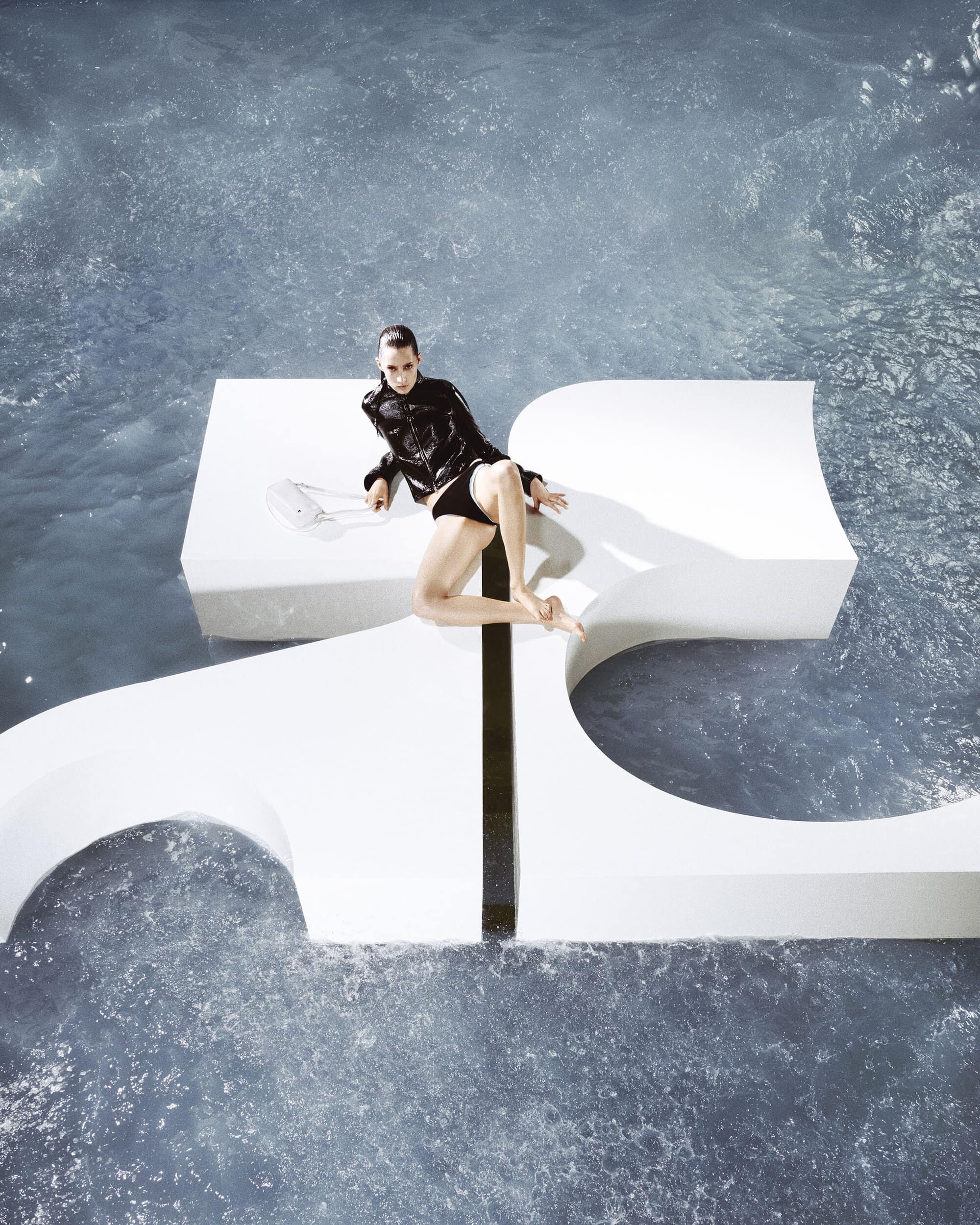 Model seated on large white geometric sculptural letters in shimmering water for Courreges digital beachwear campaign