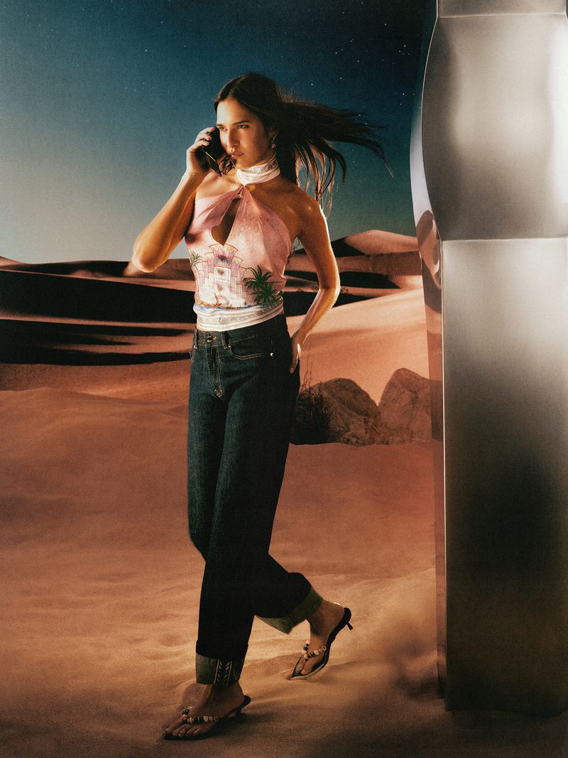 Model talking on a smartphone wearing a patterned pink halter top and dark jeans in a desert landscape at dusk