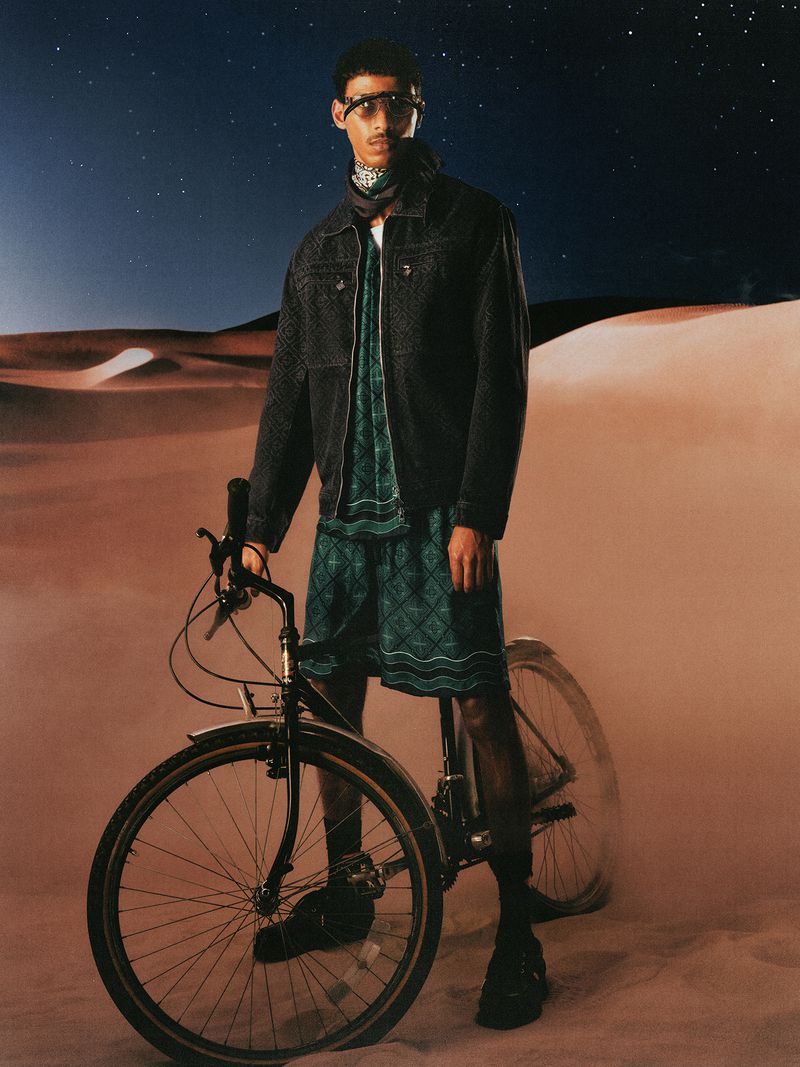 Model standing with bicycle wearing a dark jacket and patterned green shorts against a sandy desert and night sky