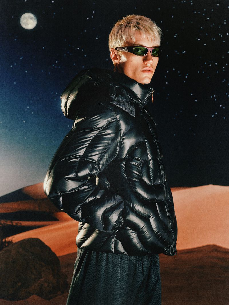Man wearing a glossy black puffer jacket and sunglasses against a starry night desert landscape