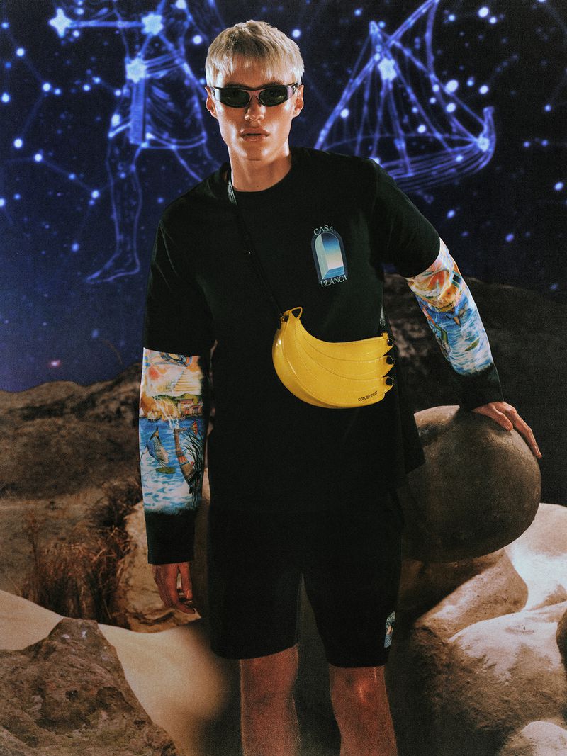 Model wearing dark sunglasses and black long-sleeve graphic tee with a yellow banana crossbody bag in a celestial landscape