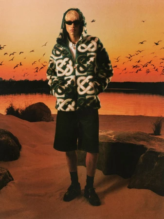 Model in patterned green and white hoodie and dark shorts standing on a sandy beach at sunset for Casablanca Pre-Fall 26 campaign.