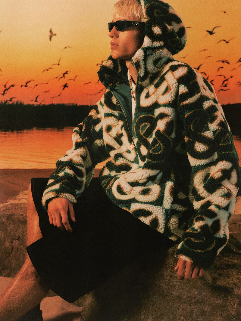 Model wearing a patterned fleece hoodie and dark shorts posed against a warm sunset sky backdrop with birds.