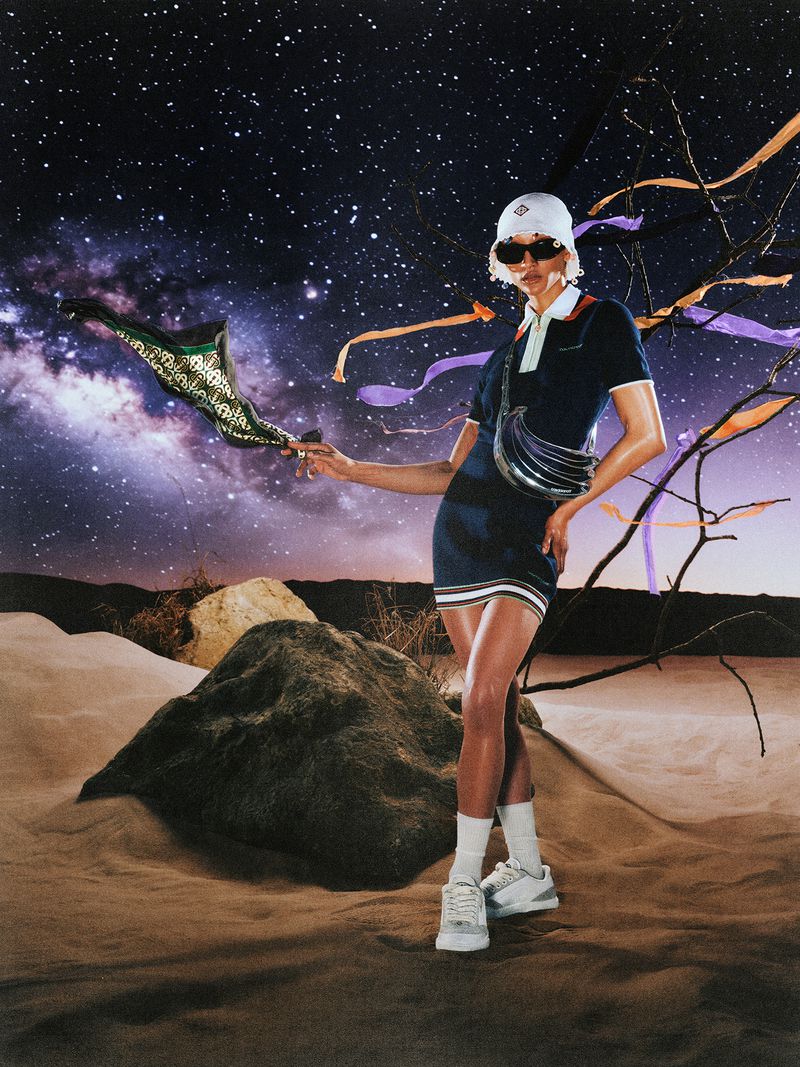 Model wearing a dark dress, white bucket hat, and sneakers standing in a desert landscape against a starry night sky backdrop.