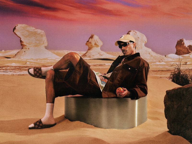 A man wearing sunglasses, a cap, and brown jacket lounging on a metallic platform set in a surreal desert landscape.