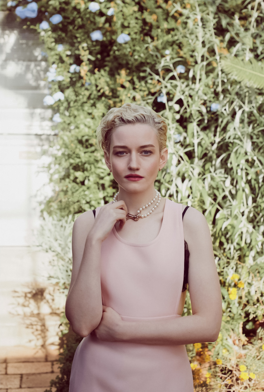 Portrait of Julia Garner wearing a pink dress and pearl necklace outdoors against a lush green garden wall.