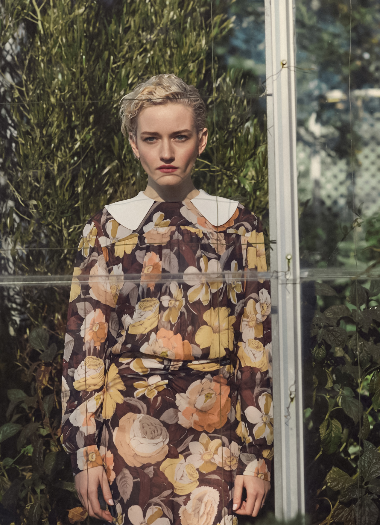 Portrait of a blonde woman in a floral dress with a white collar, standing in front of greenery and a greenhouse-like structure.