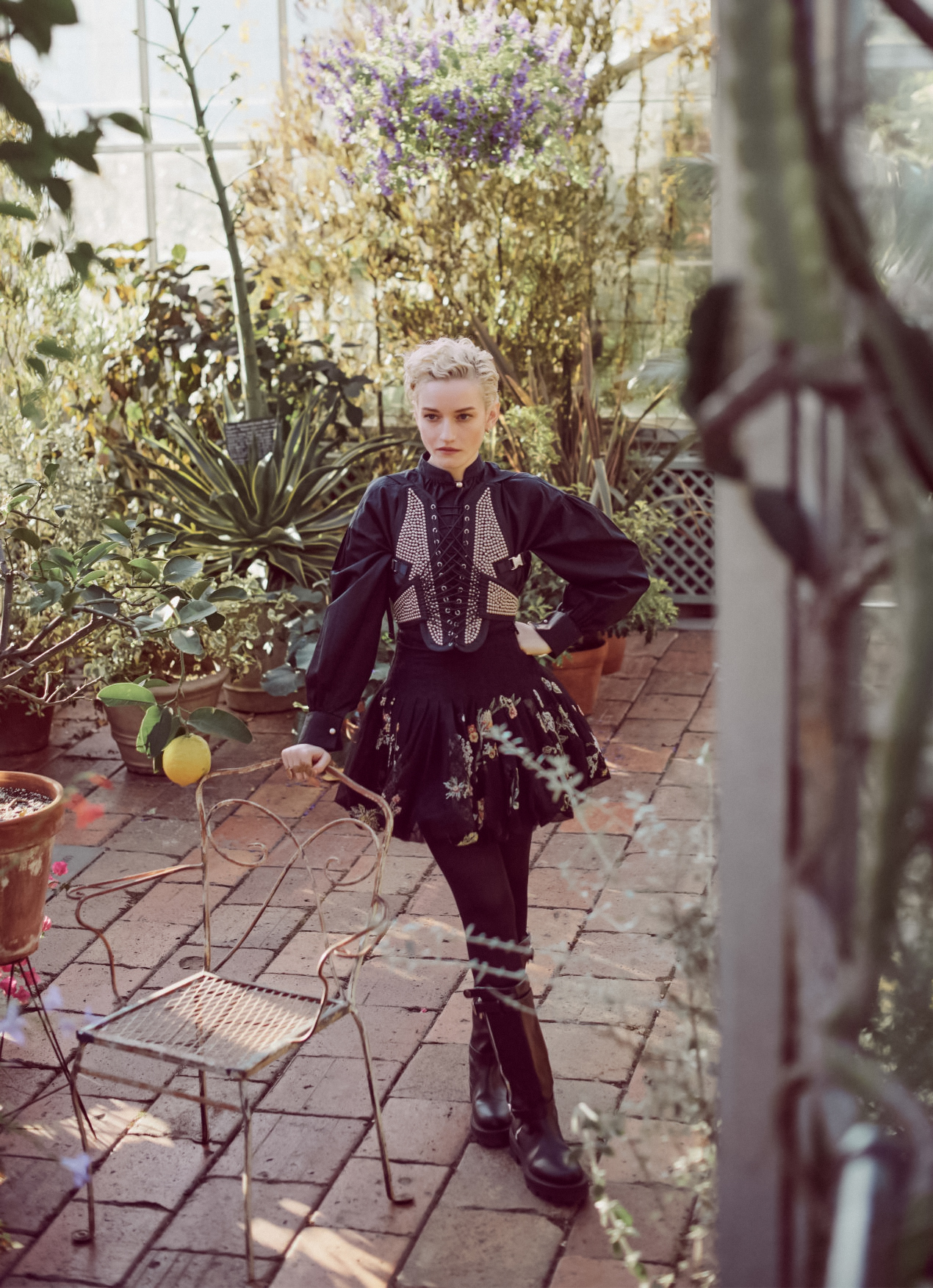 Fashionable woman in a black patterned dress stands near a metal chair on a brick patio with greenery and flowers in the background.