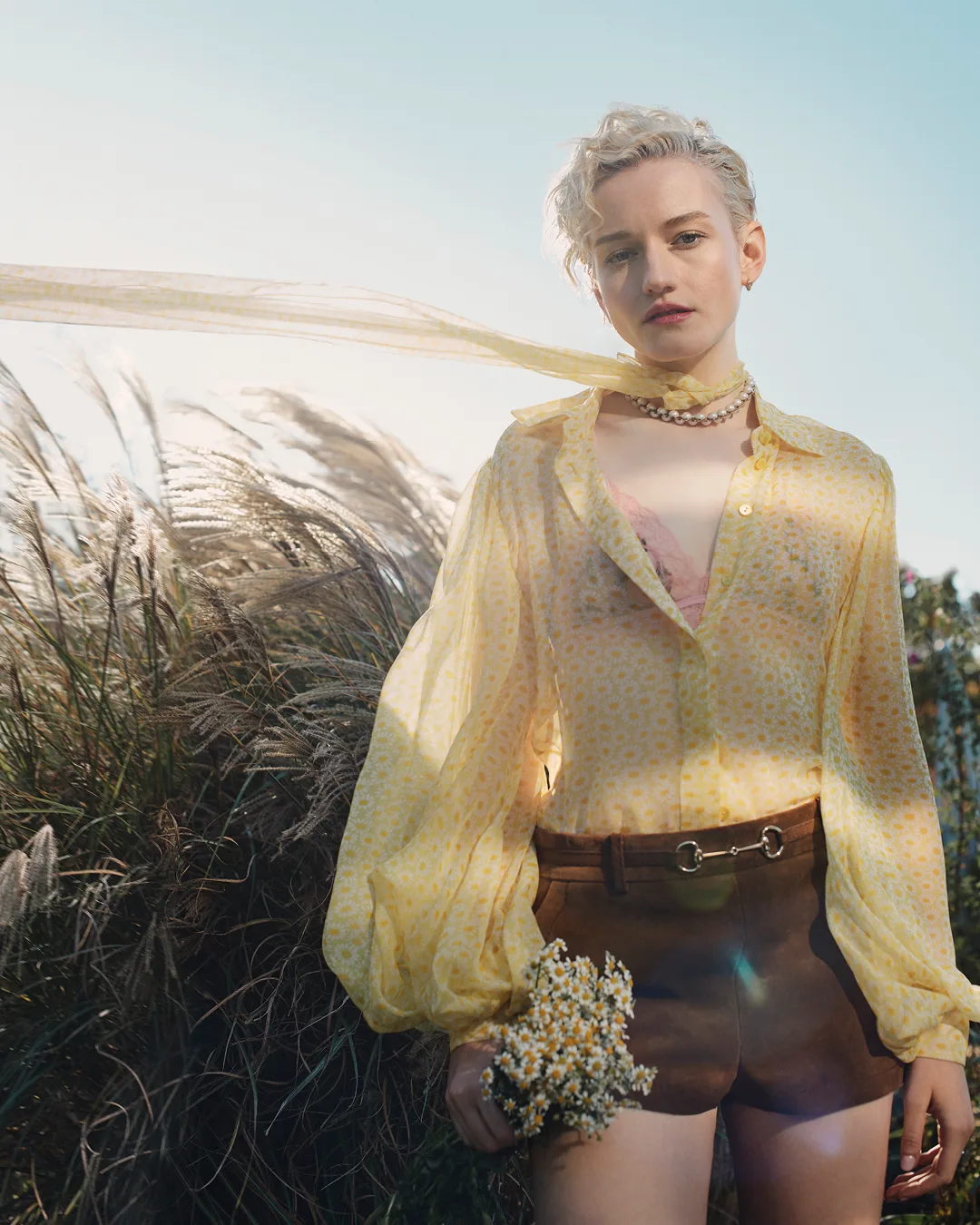 Blonde woman wearing a yellow sheer blouse and brown shorts holding wildflowers outdoors under a clear sky