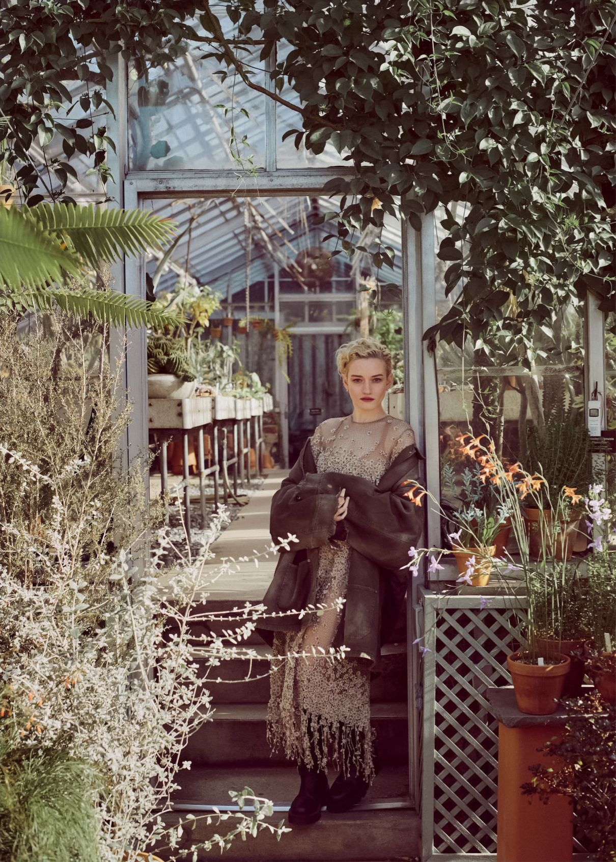 Blonde woman poses on the steps of a greenhouse, wearing a vintage-style dress and jacket surrounded by lush greenery and plants.