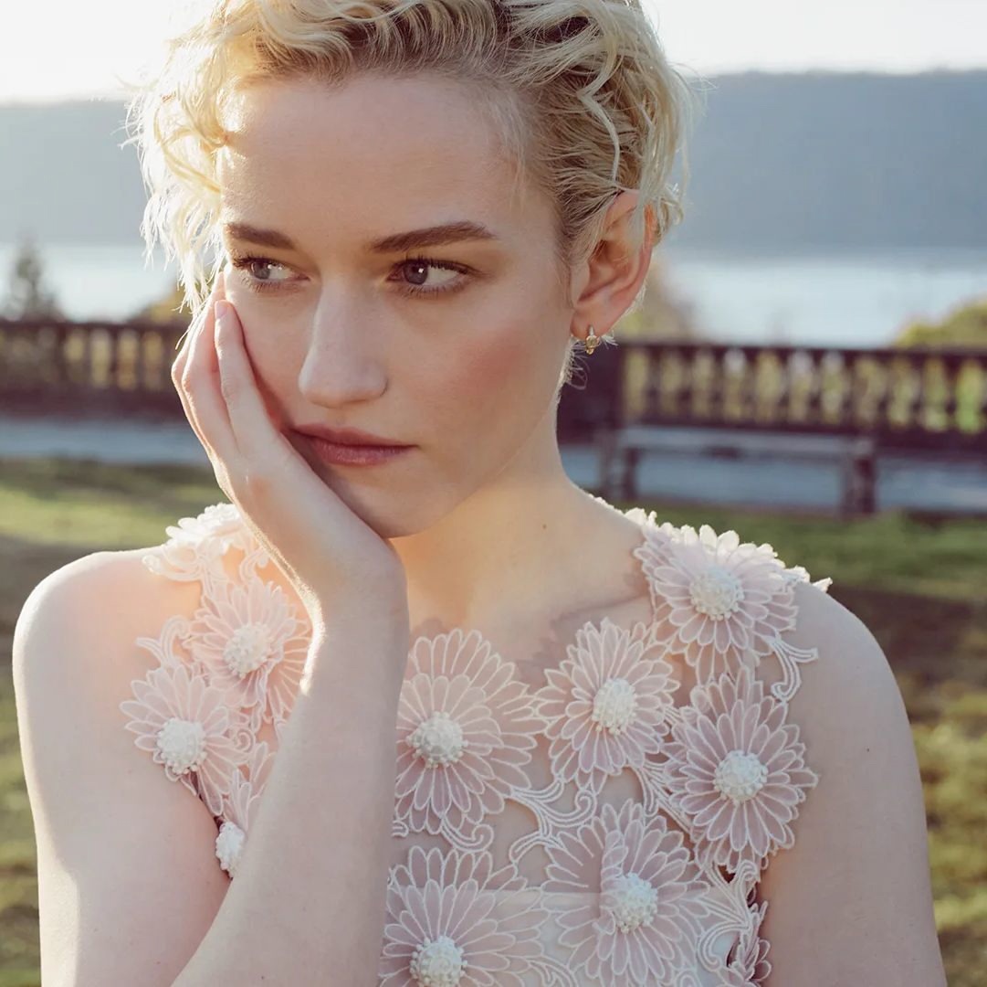 Close-up portrait of a blonde woman with her hand on her face, wearing a floral-patterned dress, and standing outside in natural lighting.