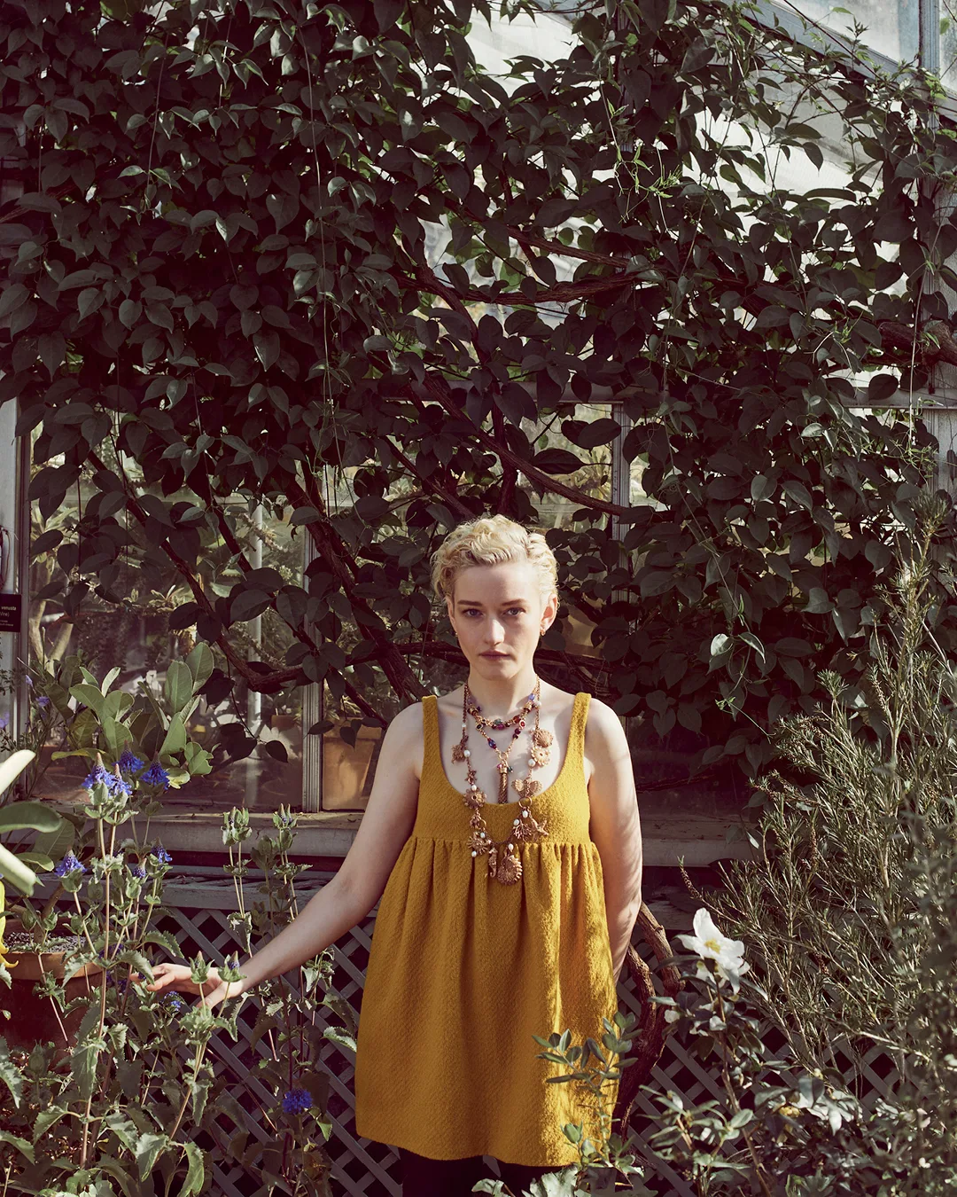 Woman in mustard dress stands in greenhouse surrounded by plants. She wears a necklace and has short, blonde hair.
