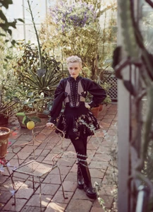 Julia Garner wearing a black dress with intricate bodice details standing inside a sunlit greenhouse