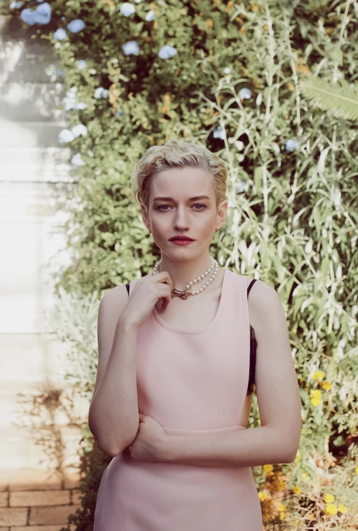 Portrait of Julia Garner wearing a pink dress and pearl necklace outdoors against a lush green garden wall.