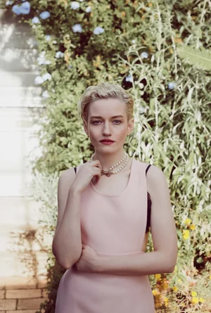 Portrait of Julia Garner wearing a pink dress and pearl necklace outdoors against a lush green garden wall.