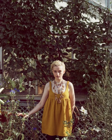 Julia Garner wearing a mustard dress and layered beaded necklace standing among lush green plants in a greenhouse
