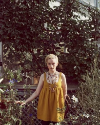 Julia Garner wearing a mustard dress and layered beaded necklace standing among lush green plants in a greenhouse