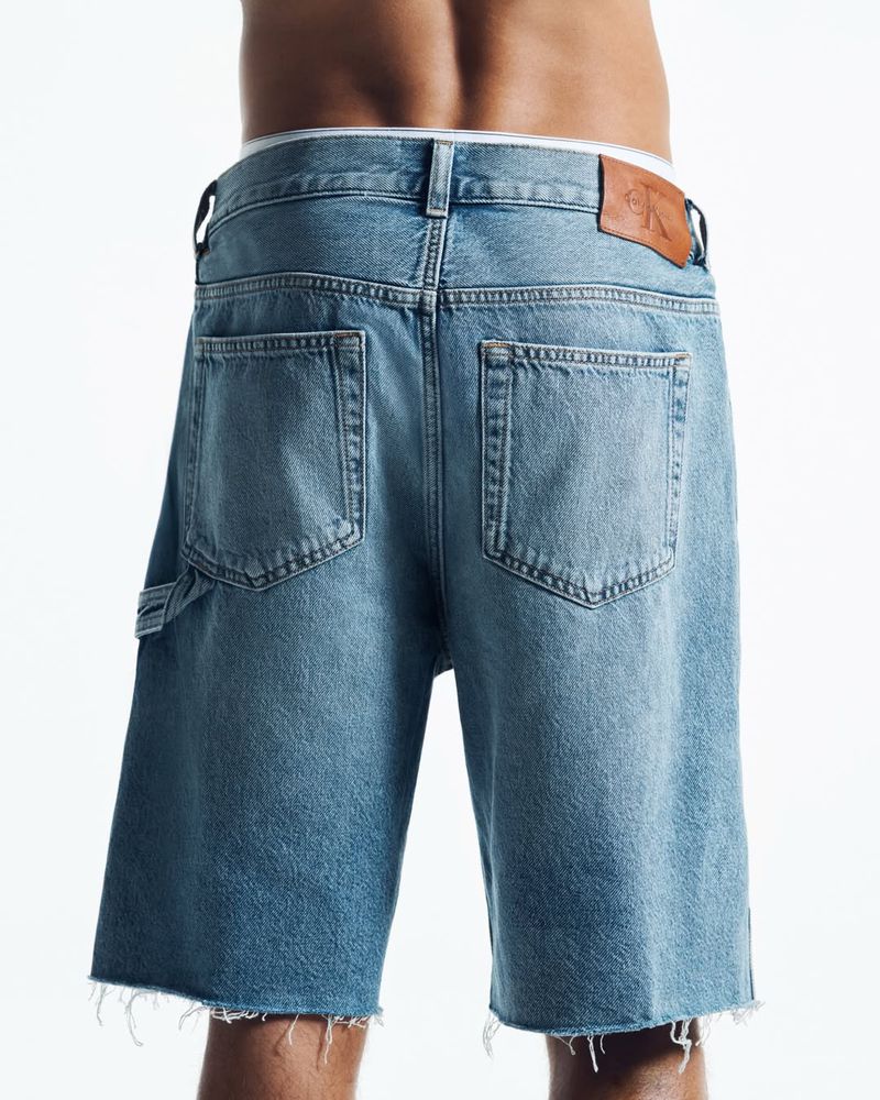 Rear view of a person wearing blue denim shorts and branded underwear waistband against a white background.
