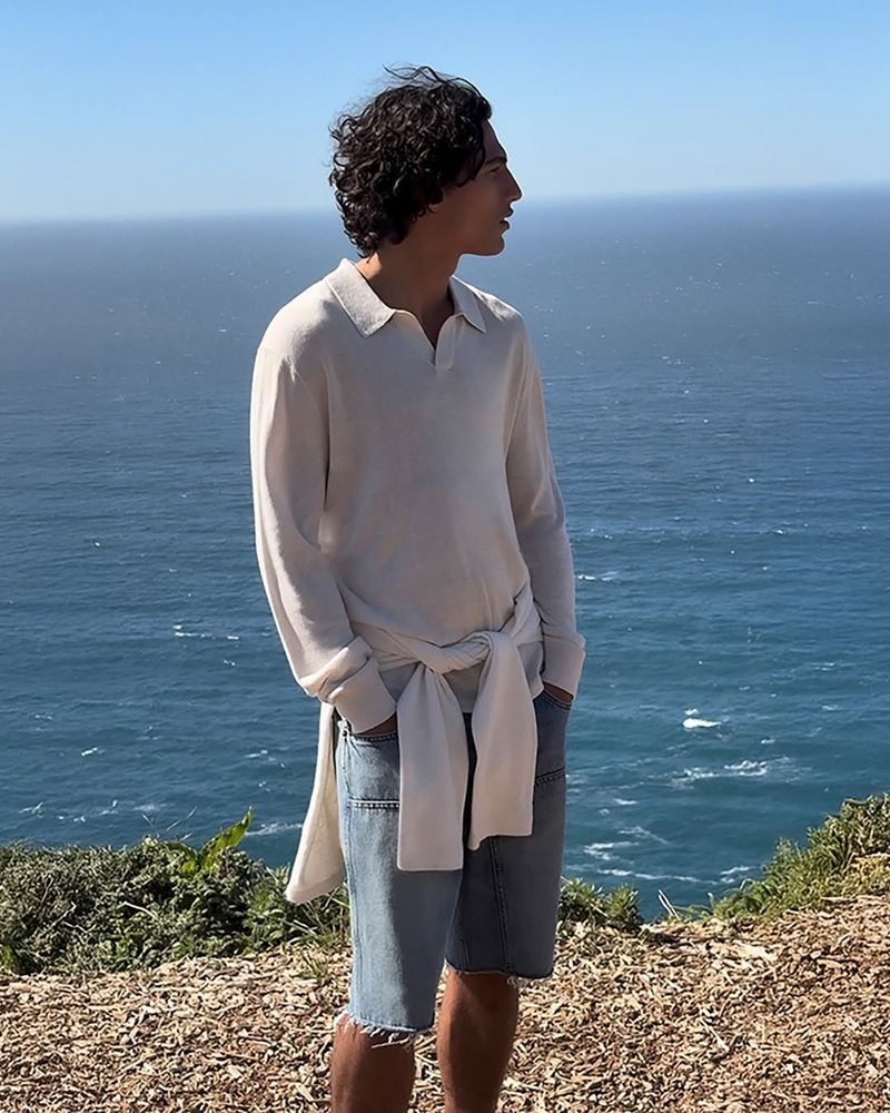 A man wearing a cream polo shirt and denim shorts standing against an ocean view backdrop with a sweater tied around his waist.