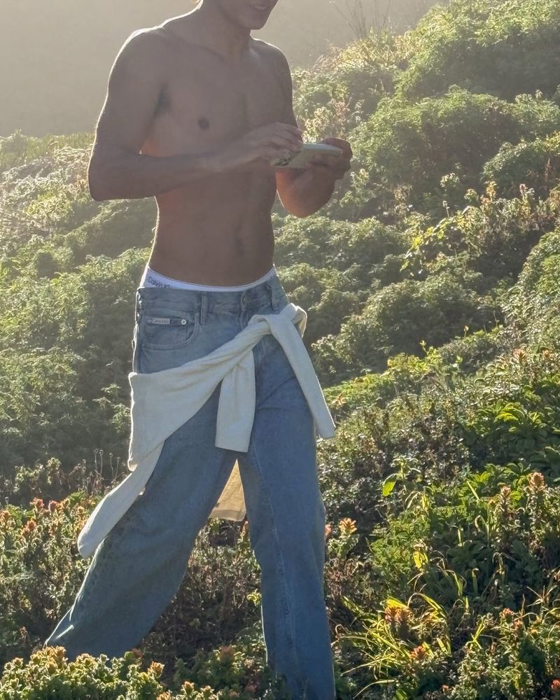 Shirtless man wearing blue jeans outdoors with a white sweater tied around his waist in natural daylight
