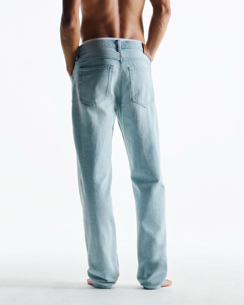 Rear view of a shirtless adult man in light blue jeans standing on a white surface in bright studio lighting; fashion and apparel