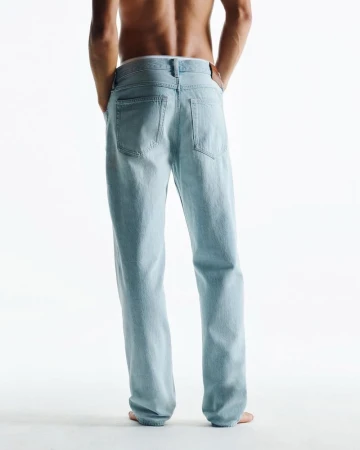 Rear view of a shirtless adult man in light blue jeans standing on a white surface in bright studio lighting; fashion and apparel