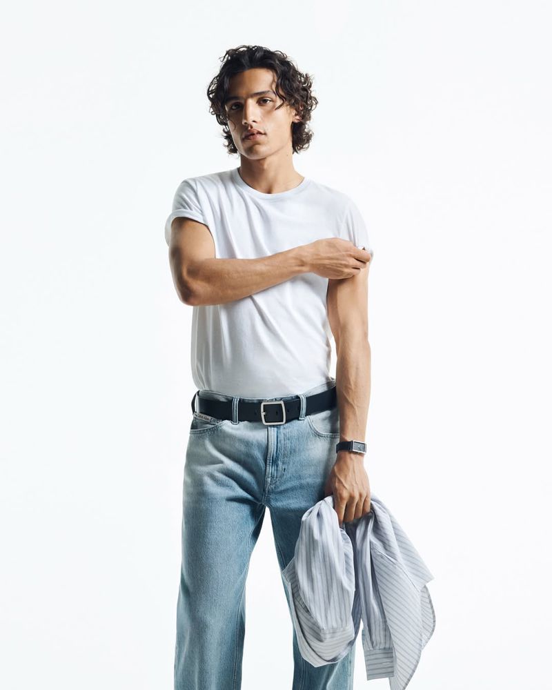 Male model wearing a white t-shirt and blue jeans holding a striped shirt against a plain white background